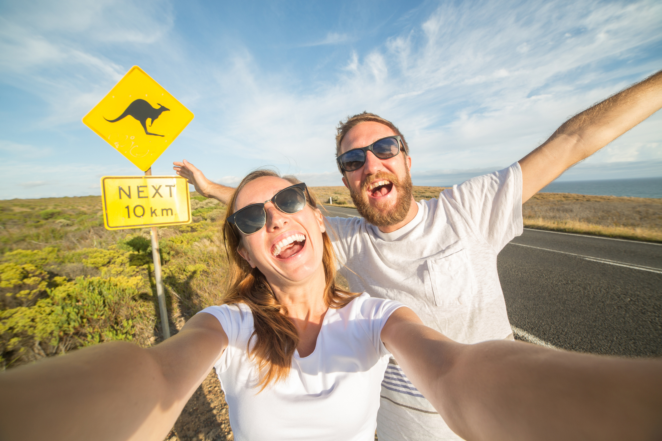 Cheering young couple take a selfie portrait on the road standing next to a kangaroo warning sign, after arriving for a gap year in Australia.